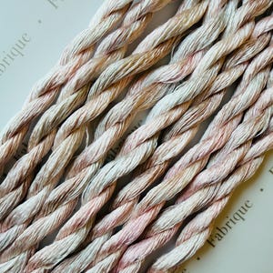 May include: Several strands of twisted embroidery thread featuring a variegated color palette of soft cream, pale pink, and muted peach tones. The threads are arranged in parallel lines, showcasing a smooth, lustrous texture and a consistent rope-like twist.
