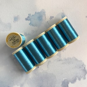 Könnte beinhalten: Sechs Spulen blauen Metallic-Garn, jede mit einem weißen Etikett mit der Aufschrift "N°40 METAL 222" und "AU CHINOIS".