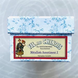 Puede incluir: Una caja azul y blanca con una etiqueta roja que dice "Fil au Chinois Ets Toulemonde Successeurs Métalisés Assortiment 2 Fabrication Française". La etiqueta también presenta una imagen de dibujos animados de una persona con un traje azul y dorado.