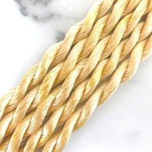 May include: Close-up of several strands of twisted, light beige cord. The cord has a slight sheen and is tightly wound, creating a textured surface. The background is a white marble-like surface.