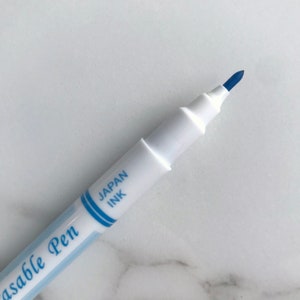 May include: Close-up of a white erasable pen with blue ink. The pen has the text "Japan Ink" and "Erasable Pen" printed on the side. The pen tip is a dark blue color.