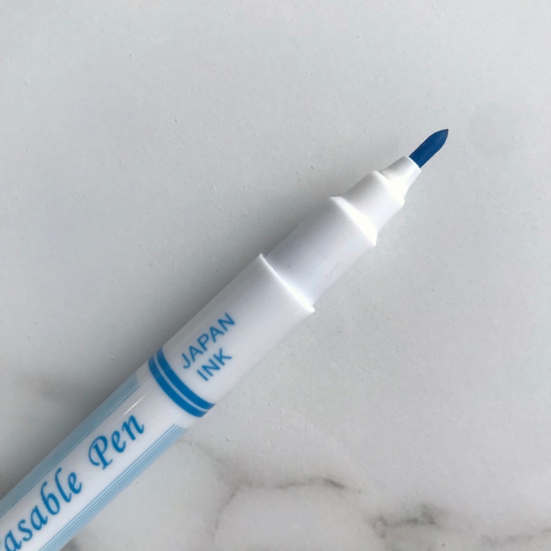May include: Close-up of a white erasable pen with blue ink. The pen has the text "Japan Ink" and "Erasable Pen" printed on the side. The pen tip is a dark blue color.