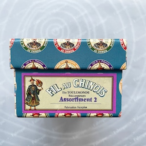 Könnte beinhalten: Eine tealfarbene Garnbox mit dem Text "FIL AU CHINOIS" und "Assortiment 2". Die Box hat ein dekoratives Muster mit einer Vintage-Illustration einer Person in traditioneller Kleidung. Der Text "Fabrication française" ist ebenfalls vorhanden.