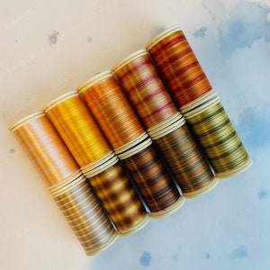 May include: A collection of spools of metallic thread in various shades of gold, copper, and bronze. The threads are wound on white plastic spools, creating a gradient of colors. The threads are arranged diagonally, showcasing their shimmering quality.
