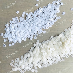 May include: Two piles of white flower-shaped sequins. The sequins are scattered on a light brown wooden surface.