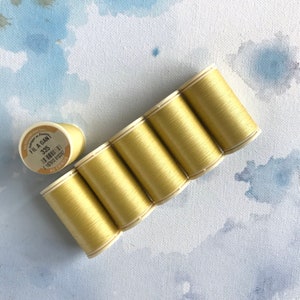 May include: Six spools of pale yellow thread, arranged in a row. The label on one spool reads "Fil a Gan 335".