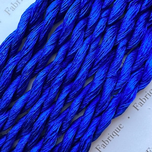 Pode incluir: Close-up de linha de bordar azul vibrante, torcida em uma estrutura semelhante a uma corda. Os fios têm um brilho brilhante, refletindo a luz. O fundo é uma superfície branca com a palavra "Fabrique" impressa.