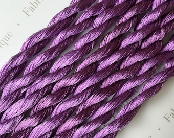 May include: A collection of metallic purple twisted cords arranged in parallel lines. The cords feature a high-sheen finish with deep violet and bright lavender tones. The word Fabrique is partially visible in the background in a light gray font.