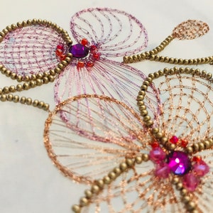 May include: Close-up of a delicate embroidered design featuring two pink and white flowers with gold beads and sparkling purple rhinestones.