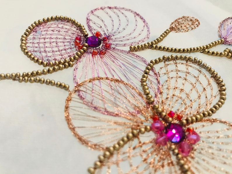 May include: Close-up of a delicate embroidered design featuring two pink and white flowers with gold beads and sparkling purple rhinestones.