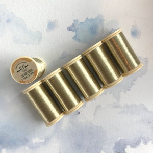 May include: Several spools of metallic gold thread are arranged diagonally on a white surface with blue watercolor accents. One spool is separate, while the others are grouped together. The label on the spool reads "METAL 102".