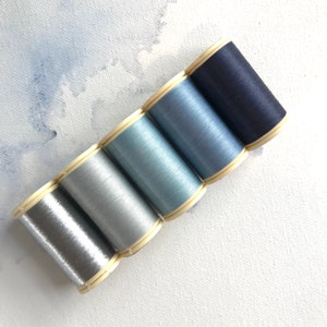 May include: Six spools of thread in shades of blue and silver. The spools are arranged in a gradient from dark blue to silver.