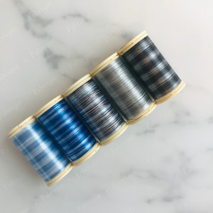 May include: Five spools of metallic thread in shades of blue, silver, and black. The thread is wound on cream-coloured spools. The spools are arranged diagonally against a white and grey marble background. The thread appears to be for sewing or embroidery.