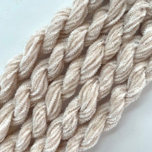 May include: Close-up of beige braided yarn. The yarn has a soft, velvety texture and is tightly twisted, creating a rope-like appearance. The color is a light, neutral beige, suitable for various crafting projects.