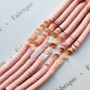 May include: Close-up of several strands of pale pink sequins, each strung together. Some strands have iridescent sequins interspersed. The sequins are flat and round, and the background is a white surface with the word "Fabrique" printed on it.