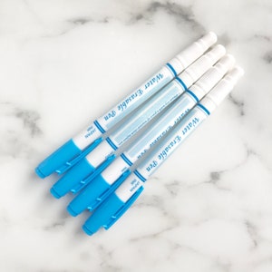 May include: Four water erasable pens with blue barrels and white caps are arranged diagonally on a marble surface. Each pen has the text "Water Erasable Pen" and "JAPAN INK" printed on it. The pens are designed for use with water.