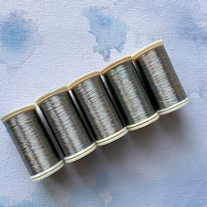 May include: Six spools of metallic thread in a silver and green color. The thread is wound around white spools.