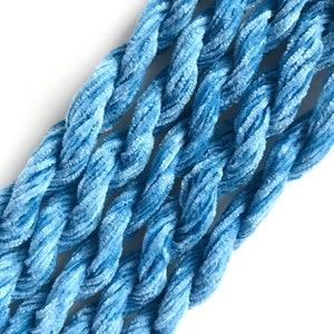 May include: Close-up of several strands of light blue, twisted velvet yarn. The yarn has a soft, plush texture and is arranged diagonally against a white background. The color is a muted, pastel blue, ideal for crafting projects.