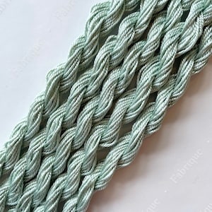 Puede incluir: Primer plano de varios carretes de hilo de bordar verde pálido. El hilo está dispuesto en forma de cruz, destacando el color suave y apagado.