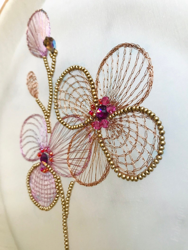 May include: A delicate floral embroidery design featuring pink and gold flowers with sparkling pink and purple rhinestones. The embroidery is on a white fabric background.