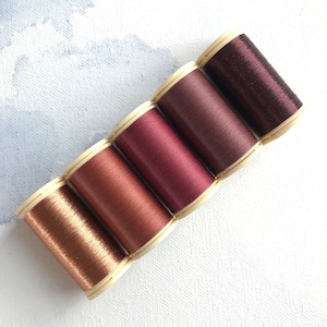 May include: Five spools of thread in shades of brown and burgundy. The spools are arranged in a gradient from light brown to dark burgundy.