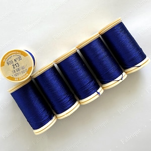 May include: A collection of navy blue silk thread spools. The spools are arranged diagonally, with one spool slightly apart from the others. The labels read "SOIE N°30 213 AU CHINOIS". The thread is tightly wound on the spools.
