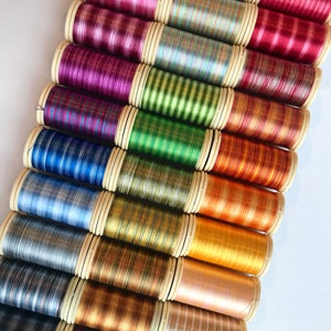 Peut inclure: Une collection de bobines de fil colorées, disposées en diagonale. Les fils sont de différentes nuances, notamment rose, violet, vert, bleu, or et marron. Chaque bobine a une base de couleur crème. Les fils semblent métalliques ou brillants.