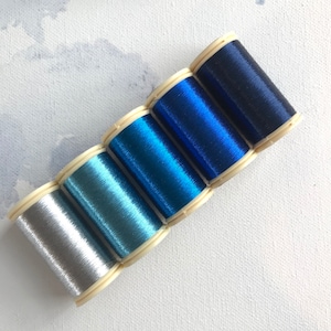 Puede incluir: Seis carretes de hilo en una gama de colores, desde plateado hasta azul oscuro. El hilo está enrollado en carretes de plástico color crema. Los colores incluyen turquesa y varias tonalidades de azul. Los hilos tienen un aspecto brillante y metálico.
