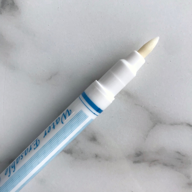May include: Close-up of a white water-erasable marker with a light beige tip. The marker has a blue stripe design and the words "Water Erasable" printed on the side. The marker is designed for fabric marking.