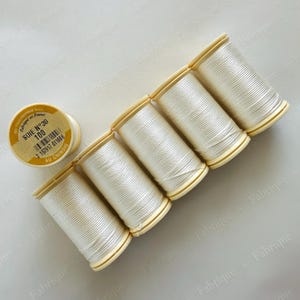 May include: A collection of ivory-colored silk thread spools. The spools are arranged diagonally, with one spool separate and the others in a row. The labels read "SOIE N°30 100 AU CHINOIS" and "Fabriqué en France".