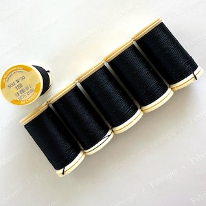 May include: A collection of black sewing thread spools with yellow labels. The labels read "SOIE N°30" and "Fabriqué en France". The spools are arranged on a white surface, showcasing the thread's dark color and texture.