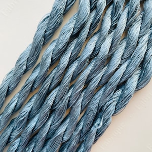 May include: Close-up of several strands of twisted, light blue embroidery floss. The floss has a slightly textured appearance and a subtle sheen. The strands are arranged diagonally across the frame, creating a pattern. The background is a plain, off-white surface.
