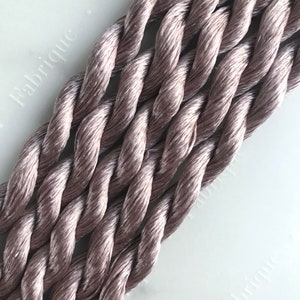 May include: Close-up of a bundle of soft, light brown, twisted embroidery floss. The floss is arranged in a diagonal pattern, with the ends of the strands visible.