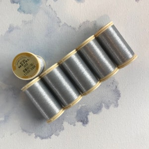 May include: Six spools of silver metallic thread, arranged in a semi-circle, with one spool of the same thread lying separately. The spool label reads "N°40 Metal 322".