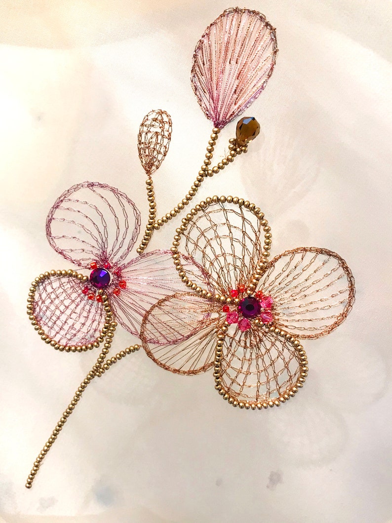 May include: A delicate gold and pink floral embroidery with purple and red rhinestones. The embroidery is on a white fabric background.