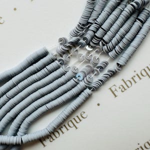 May include: Close-up of strands of gray sequins, each with a small hole in the center, strung together on clear thread. The sequins are arranged in multiple parallel lines, creating a textured surface. The background is a white surface with the word "Fabrique" printed on it.
