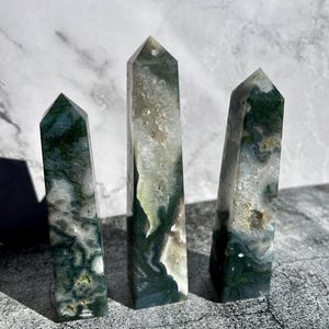 May include: Three moss agate crystal obelisks. The crystals are a mix of white, clear, and green, with a polished, smooth surface. The obelisks are of varying heights, with pointed tops. The crystals are displayed on a gray surface.