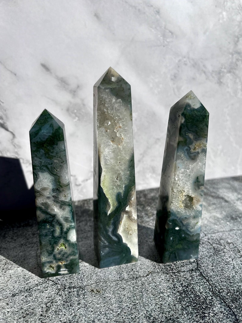 May include: Three moss agate crystal obelisks. The crystals are a mix of white, clear, and green, with a polished, smooth surface. The obelisks are of varying heights, with pointed tops. The crystals are displayed on a gray surface.