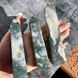 May include: Three polished moss agate crystal towers. The crystals have a mix of white, green, and gray colors. The towers are rectangular with pointed tops. The crystals are held in a hand against a gray background.