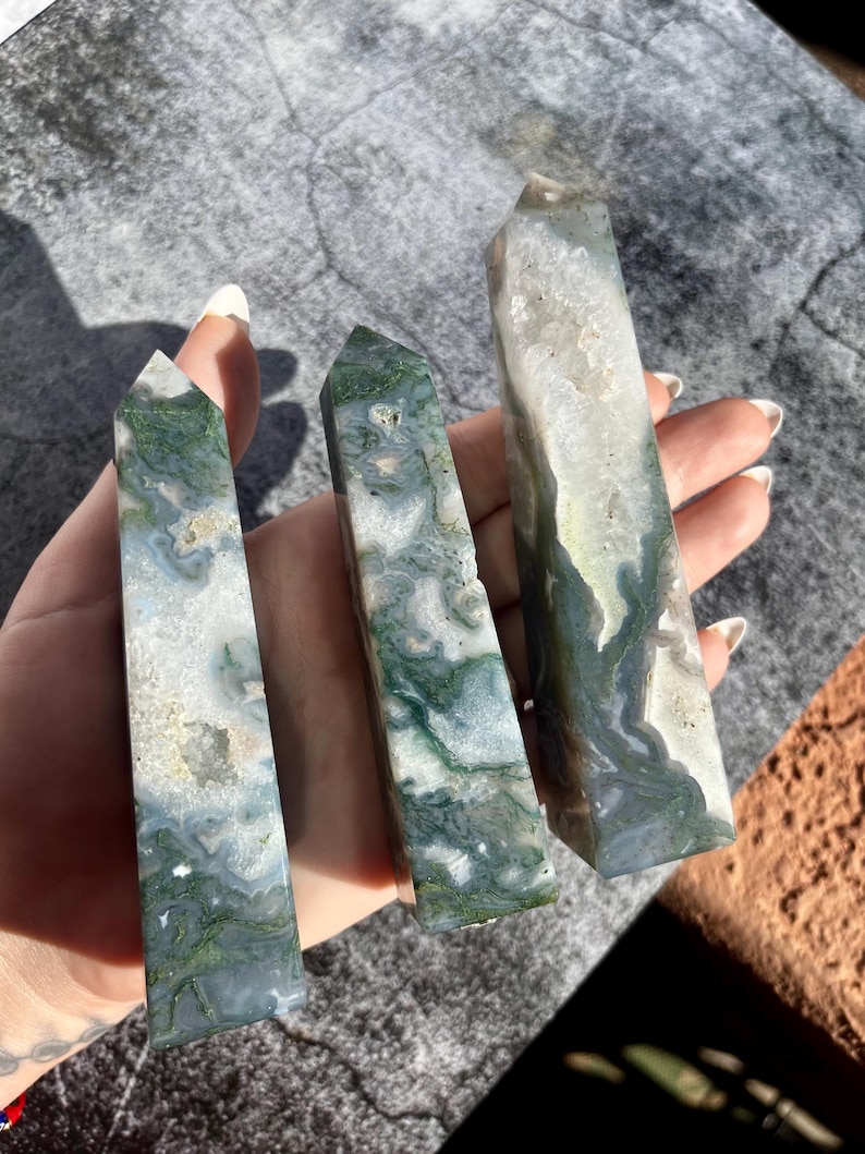 May include: Three polished moss agate crystal towers. The crystals have a mix of white, green, and gray colors. The towers are rectangular with pointed tops. The crystals are held in a hand against a gray background.
