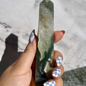 May include: A tall, rectangular crystal point with a polished surface. The crystal features a mix of clear, white, and green hues, with darker green inclusions. The crystal is held against a white and gray marble background.