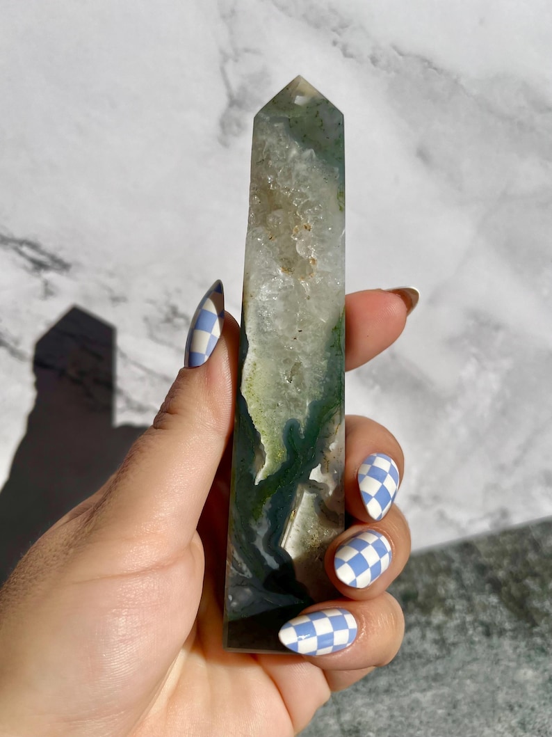 May include: A tall, rectangular crystal point with a polished surface. The crystal features a mix of clear, white, and green hues, with darker green inclusions. The crystal is held against a white and gray marble background.
