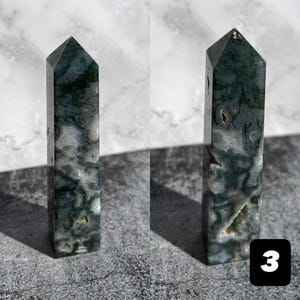 May include: Two polished moss agate obelisks. The obelisks are rectangular with a pointed top. The stone has a mix of white, green, and gray colors. The obelisks are standing on a gray surface. A black square with the number 3 is in the bottom right corner.
