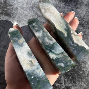 May include: Three polished moss agate obelisks. The stones feature a mix of white, green, and gray colors with unique patterns. The obelisks are rectangular with a pointed top. The stones are held in a hand.