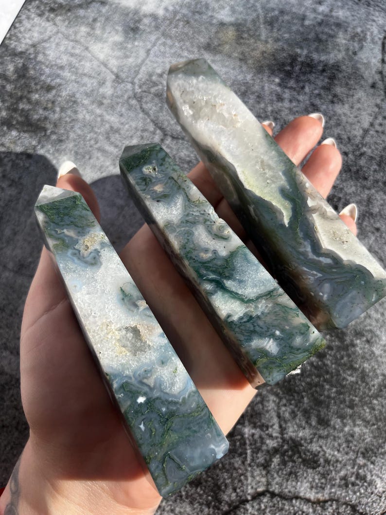 May include: Three polished moss agate obelisks. The stones feature a mix of white, green, and gray colors with unique patterns. The obelisks are rectangular with a pointed top. The stones are held in a hand.