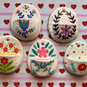 May include: Five oval-shaped ceramic wall pockets with hand-painted floral and geometric designs. Each piece features a small circular hole at the top for hanging and a semi-circular pocket at the bottom, decorated with vibrant colours like blue, pink, red, and green.