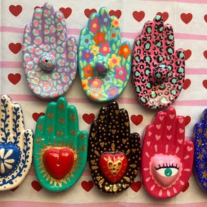 May include: A collection of nine hand-shaped ceramic incense holders. Each holder features unique designs, including floral patterns, leopard print, and hearts. Colors include pink, blue, green, and gold. The holders are arranged on a pink and white striped surface with red heart accents.