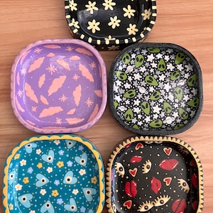 May include: Five square-shaped decorative trays with painted designs. The trays are black, purple, teal, and black with gold trim. The designs include flowers, clouds, hearts, eyes, and lips.