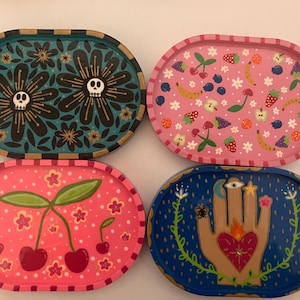 May include: Four oval decorative trays with colorful designs. One features black flowers with skulls, another has pink with fruit, a third has cherries, and the last has a hand with an eye and heart.
