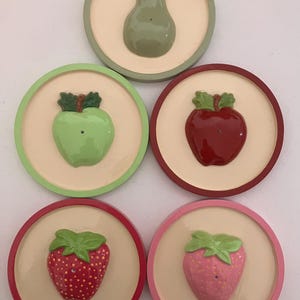 May include: A set of five circular ceramic coasters, each featuring a raised fruit design in the center. The collection includes a green pear, a light green apple, a red apple, a red strawberry, and a pink strawberry, all on cream bases.